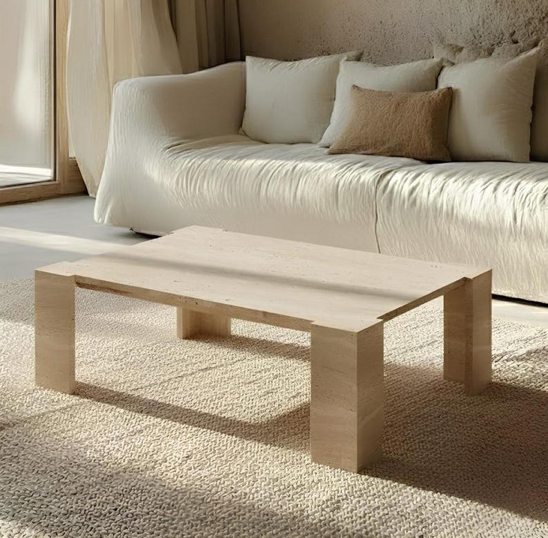 May include: A light-colored rectangular coffee table with a minimalist design. The table has a solid, blocky construction with four thick legs. It sits on a textured, light-colored rug, with a white sofa and pillows in the background.
