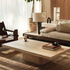 Pode incluir: Uma mesa de centro retangular de cor clara com uma superfície texturizada está sobre um tapete tecido. Uma pilha de livros, uma tigela pequena e um copo decorativo estão sobre a mesa. Um sofá, uma lâmpada e uma cadeira estão ao fundo, criando um ambiente acolhedor na sala de estar.