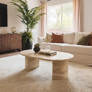 May include: An oval coffee table crafted from beige travertine stone with a smooth top surface and two curved, solid pedestal bases. The table features the natural porous texture and light tan veining characteristic of travertine stone construction.