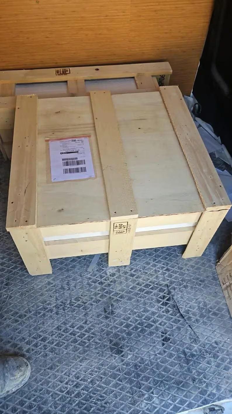 May include: A large, rectangular wooden crate with a light brown finish. The crate has a lid and is reinforced with vertical and horizontal wooden slats. A shipping label is affixed to the top.