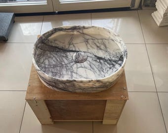 May include: Oval marble sink with a white and gray veined pattern. The sink is set on a wooden stand. The sink has a central drain hole. The overall design is modern and elegant, suitable for a bathroom.