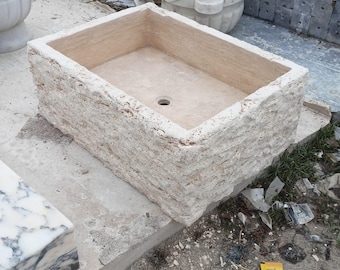 Op de afbeelding: Een rechthoekige, beige stenen wastafel met een ruwe, getextureerde buitenkant. De wastafel heeft een gladde binnenkant en een centraal afvoergat. Andere stenen wastafels en architectonische elementen zijn zichtbaar op de achtergrond.
