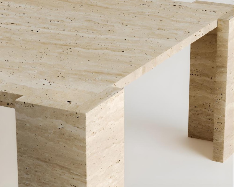 May include: A rectangular travertine table with a light beige color and dark speckles. The table has a minimalist design with a thick top and two supporting legs. The table's dimensions are approximately 60 inches long, 30 inches wide, and 16 inches high.