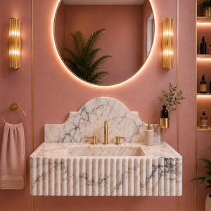 May include: A wall-mounted white marble sink with a fluted front and a scalloped backsplash, featuring gold hardware. Above the sink is a round, backlit mirror flanked by two vertical gold wall sconces against a dusty rose wall.