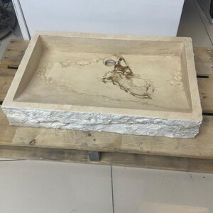 Puede incluir: Fregadero rectangular de piedra con interior beige claro y exterior rugoso y texturizado. El fregadero tiene un solo orificio de desagüe y está colocado sobre un palé de madera. El diseño del fregadero es sencillo y moderno, adecuado para un baño.
