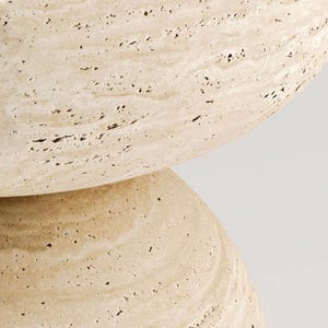 May include: Close-up of a beige stone object with a rounded shape. The surface has a textured appearance with small dark spots and subtle veining. The object appears to be made of natural stone.
