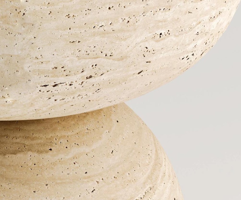 May include: Close-up of a beige stone object with a rounded shape. The surface has a textured appearance with small dark spots and subtle veining. The object appears to be made of natural stone.