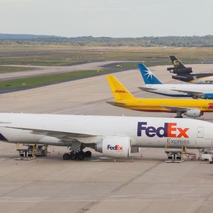 May include: A fleet of cargo airplanes parked on a tarmac. The lead plane is white with a purple tail and the FedEx logo. Other planes include DHL, UPS, and Star Air. Ground support equipment is visible.
