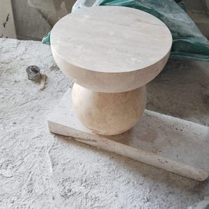 May include: A light beige stone side table with a round top and a spherical base. The table is made of a natural stone material and sits on a rectangular stone base. The background shows other stone pieces.