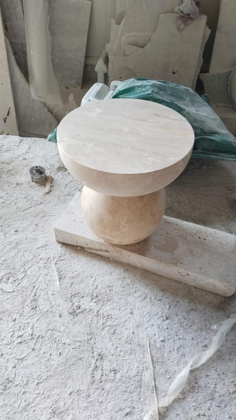 May include: A light beige stone side table with a round top and a spherical base. The table is made of a natural stone material and sits on a rectangular stone base. The background shows other stone pieces.
