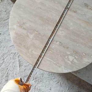 May include: A round, light beige marble table with a tape measure laid across its surface. The table's diameter is being measured. The person measuring is wearing work gloves and white sneakers. The table is on a concrete surface.