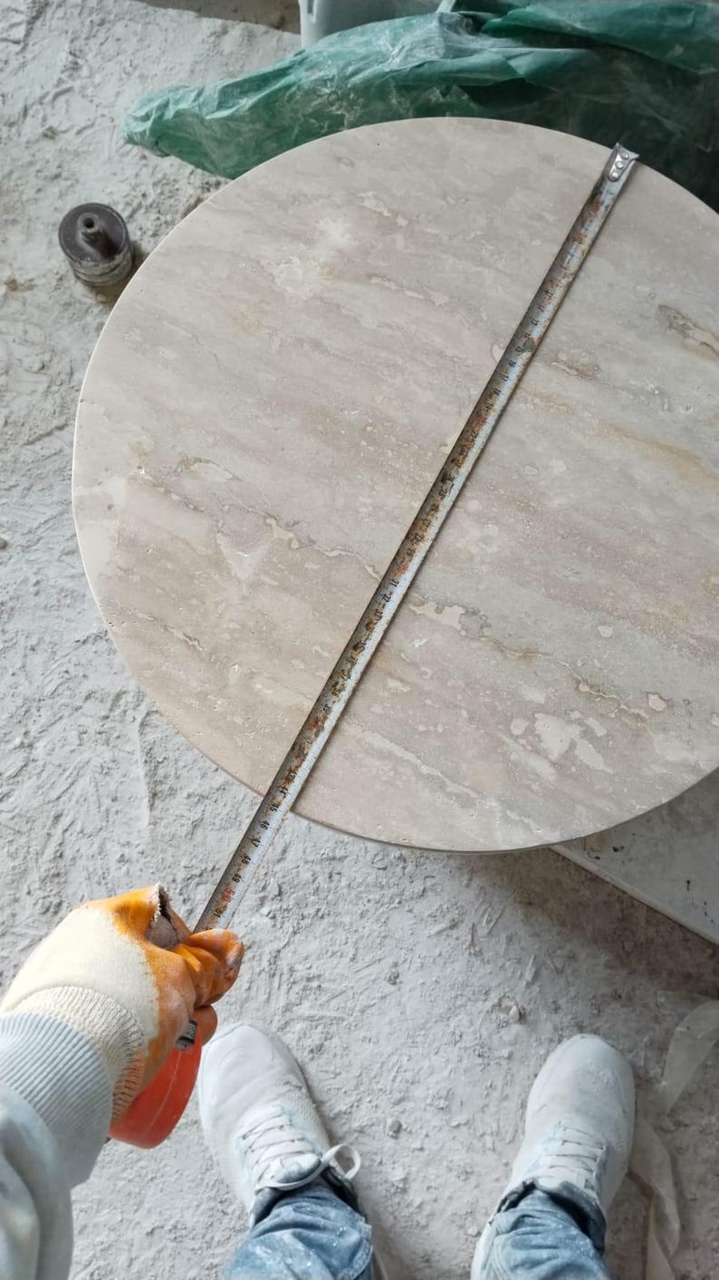 May include: A round, light beige marble table with a tape measure laid across its surface. The table's diameter is being measured. The person measuring is wearing work gloves and white sneakers. The table is on a concrete surface.