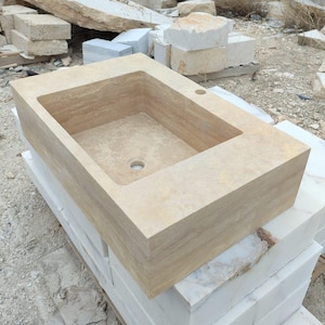 May include: A rectangular, beige stone sink with a recessed basin and a single faucet hole. The sink is made of a light-colored stone and sits atop a stack of white marble blocks. The overall theme is bathroom fixtures.