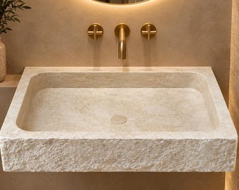 May include: A rectangular stone sink with a rough, chiseled exterior texture and a smooth interior basin. The sink is mounted to a wall beneath a gold-toned faucet with two cross-shaped handles and a central spout, positioned below a round mirror.