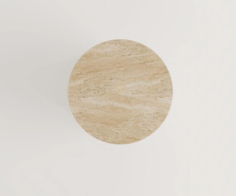 May include: Overhead view of a round, beige travertine stone table. The table has a natural, textured surface with subtle variations in color and pattern. The table is set against a white background.
