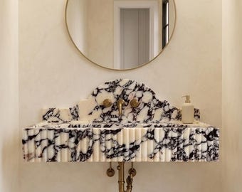 May include: A marble vanity with black and white veining, featuring a scalloped edge and brass fixtures. A round gold-framed mirror hangs above the sink, with a neutral-toned wall and a white door in the background. A soap dispenser sits on the vanity.
