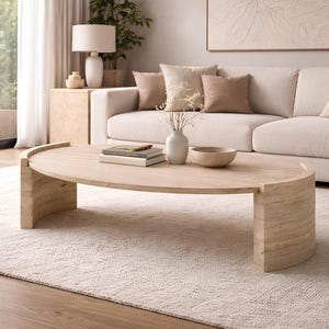 May include: A light-coloured, oval coffee table with a rounded base. The table is made of wood and has a natural finish. Books and a wooden bowl sit on top. The table is in a living room with a beige sofa and rug. The table is approximately 120 cm long.