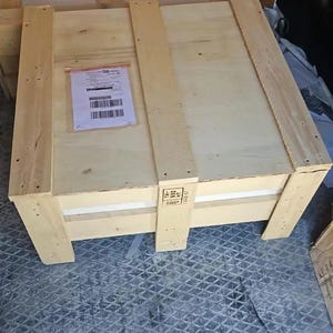 May include: A large, rectangular wooden crate with a light-colored finish. The crate is constructed with thick wooden planks and reinforced with additional wooden supports. A shipping label is affixed to the top.