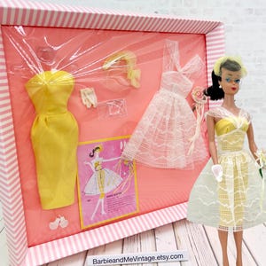 May include: A vintage Barbie doll wearing a yellow and white dress, with a matching hat and gloves. The doll is next to a framed display of vintage Barbie fashions, including a yellow dress, a white lace dress, and accessories. The frame has a pink and white striped border.