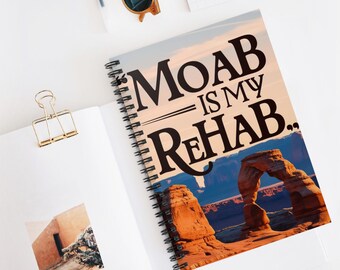 May include: A spiral notebook with a colorful image of a desert landscape featuring a natural stone arch. The text "MOAB IS MY REHAB." is printed in black on the front cover.