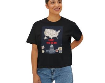 Può includere: T-shirt nera con una grafica raffigurante una mappa degli Stati Uniti, la scritta "I Didn't Vote For Him" e un'illustrazione del Campidoglio degli Stati Uniti. La maglietta è indossata da una persona con jeans blu.