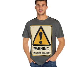 Op de afbeelding: Een grijs t-shirt met een geel waarschuwingsbord met een zwart uitroepteken en de tekst "WARNING MAY CONTAIN DAD JOKES". Het bord staat op een beige achtergrond.