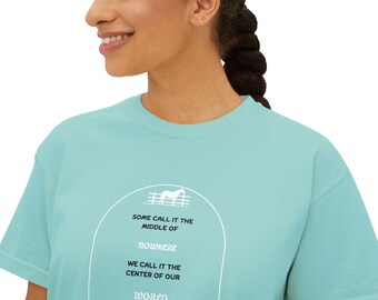 Può includere: Una t-shirt blu chiaro con una grafica bianca di una lapide con il testo "Some call it the middle of nowhere. We call it the center of our world."