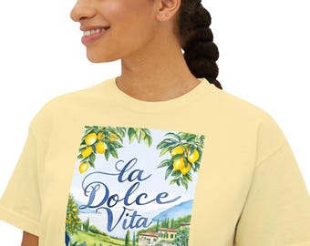 May include: A light yellow t-shirt featuring a watercolor design with the text "La Dolce Vita." The design includes lemons, green foliage, and a scenic Italian landscape with houses and rolling hills. The shirt is a comfortable, casual style.
