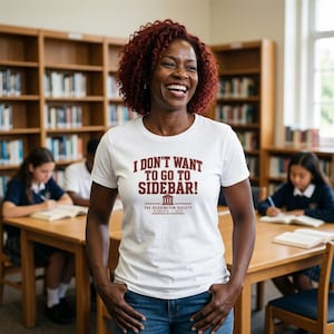 May include: A white short-sleeved t-shirt featuring the maroon text I DON'T WANT TO GO TO SIDEBAR! above a small building icon and the text THE REDDINGTON SOCIETY FITWORTH 2025. The shirt is worn with blue denim jeans in a library setting.
