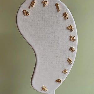 May include: Gold-colored stud earrings featuring various zodiac symbols, each with a small, sparkling accent. The earrings are displayed on a white, curved earring holder against a soft green background. The earrings are small and delicate.