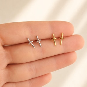 May include: A close-up of two pairs of small stud earrings. One pair is silver-colored, and the other is gold-colored. Each earring is shaped like a miniature sword or dagger, with a detailed hilt and blade.