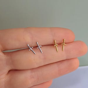 May include: Two pairs of small dagger-shaped stud earrings. One pair is silver-colored, and the other is gold-colored. The earrings are displayed on a light background. The earrings are small and delicate, suitable for everyday wear.