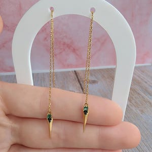 May include: Gold-coloured dangle earrings with a distinctive design. Each earring has a fine chain holding a pointed, gold-toned piece. The base of each piece is decorated with a small, dark green gemstone. The earrings are shown on a white stand.