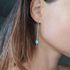 May include: A silver chain earring with a small, light blue teardrop-shaped charm. The earring is worn in the ear and hangs down. The charm has a subtle iridescent quality.
