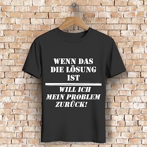 Peut inclure: T-shirt noir avec texte blanc. Le texte se lit "WENN DAS DIE LÖSUNG IST WILL ICH MEIN PROBLEM ZURÜCK!" en gras, police sans-serif. Le t-shirt est suspendu à un cintre en bois contre un mur de briques.