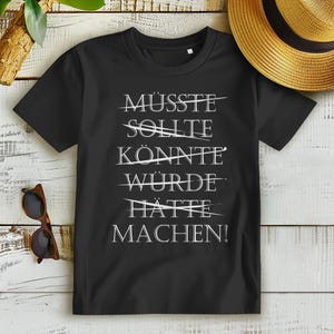May include: Black t-shirt with white text that reads "MUSSSTE, SOLLTE, KÖNNTE, WÜRDE, HATTE, MACHEN!" with lines drawn through the first five words.