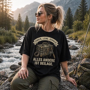 Op de afbeelding: Zwart T-shirt met een vis-thema grafisch ontwerp. Het ontwerp bevat een tacklebox, hengel en de Duitse tekst "Haken, Leine, Geduld, Alles Andere Ist Beilage." Het shirt wordt buiten gedragen.