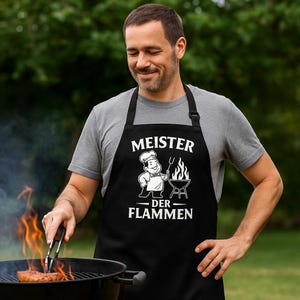 Peut inclure: Tablier noir avec l'inscription blanche "MEISTER DER FLAMMEN", un dessin de chef et un barbecue. Une personne porte le tablier en grillant de la nourriture à l'extérieur.