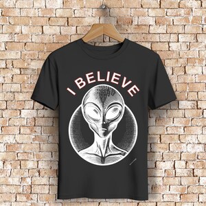 Può includere: Maglietta nera con una grafica bianca di una testa aliena in un cerchio. Il testo "I BELIEVE" è scritto in bianco sopra la testa dell'alieno.