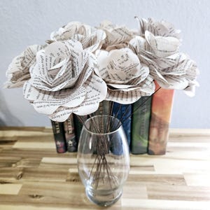 May include: A bouquet of six white paper roses made from the pages of books. The roses are arranged in a clear glass vase and are sitting on a wooden surface in front of a stack of books.