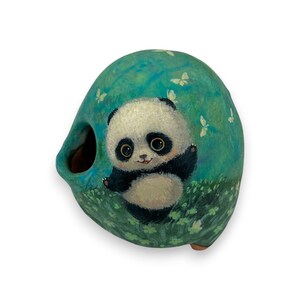 May include: A hand-painted ceramic panda bear sculpture. The panda, in black and white, is set against a teal background with butterflies and clover. The sculpture has a unique, rounded shape with an opening, suggesting a decorative item.