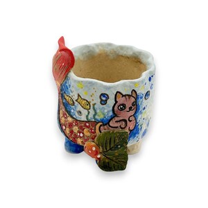 Puede incluir: Un pequeño macetero de cerámica con forma de sirena con un gato sentado en su cola. El macetero está pintado con colores azul, verde, amarillo, rojo y blanco. El gato es rosa y tiene la cara blanca. El macetero tiene una pequeña abertura en la parte superior para plantar.