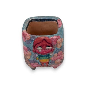 May include: A ceramic planter with a cartoon illustration of a crying character. The planter is painted with a blue background, pink flowers, and a sad character wearing a rainbow shirt. The interior is brown.