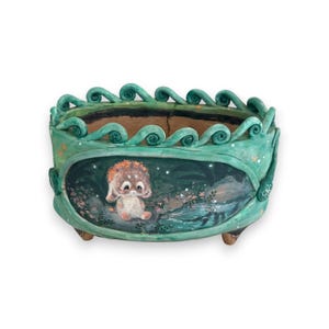 May include: A green ceramic planter with a whimsical design. The planter features a painted scene of a small animal sitting in a forest setting. The planter has a scalloped rim and three small feet.