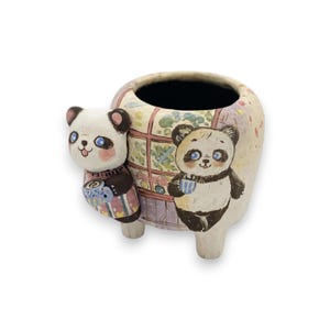May include: A ceramic planter shaped like a house with two panda bears painted on the front. The panda bears are holding cups and have black eyes and pink cheeks. The planter has a brown, black, pink, and white colour scheme.