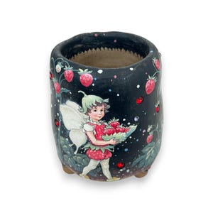 May include: A black ceramic planter featuring a fairy painting. The fairy is holding a basket of strawberries. The planter has red glitter accents and is decorated with a strawberry vine.