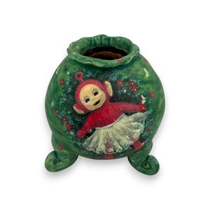 Puede incluir: Un jarrón de cerámica verde con tres patas y cuerpo redondo. El jarrón está pintado con un personaje de dibujos animados que lleva un vestido rojo y un tutú blanco.