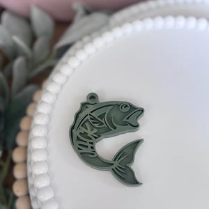 May include: A sage green fish-shaped charm with the name "JACK" cut out within the design. The charm has a hole at the top for hanging and is set against a white background.