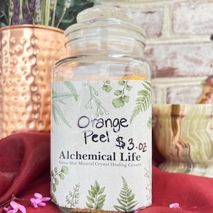 May include: A glass jar filled with orange peel powder. The label reads "Orange Peel $3.0z Alchemical Life Anna Mae Mineral Crystal Healing Center".