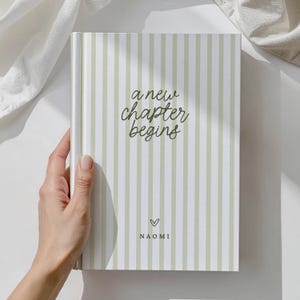 May include: A journal with a green and white striped cover. The words "a new chapter begins" are written in cursive. Below the text is a heart symbol and the name "NAOMI".